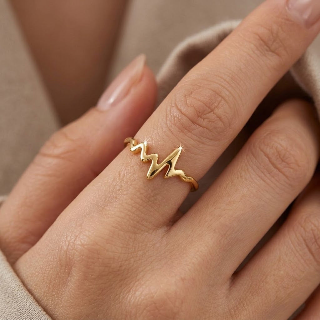 Gold ring with heart beat design on a person's finger against a neutral background