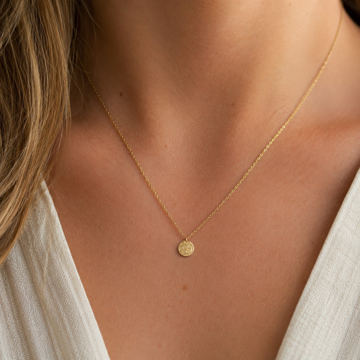 A delicate gold chain necklace with a small, round, textured gold pendant worn by a model. The necklace is shown close-up against the model's neck and collarbone, wearing a white top.