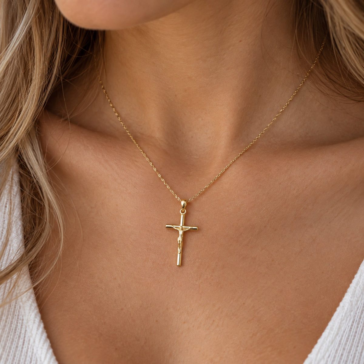 A close-up of a woman wearing a delicate gold chain necklace with a small gold crucifix pendant, styled with a white ribbed top.