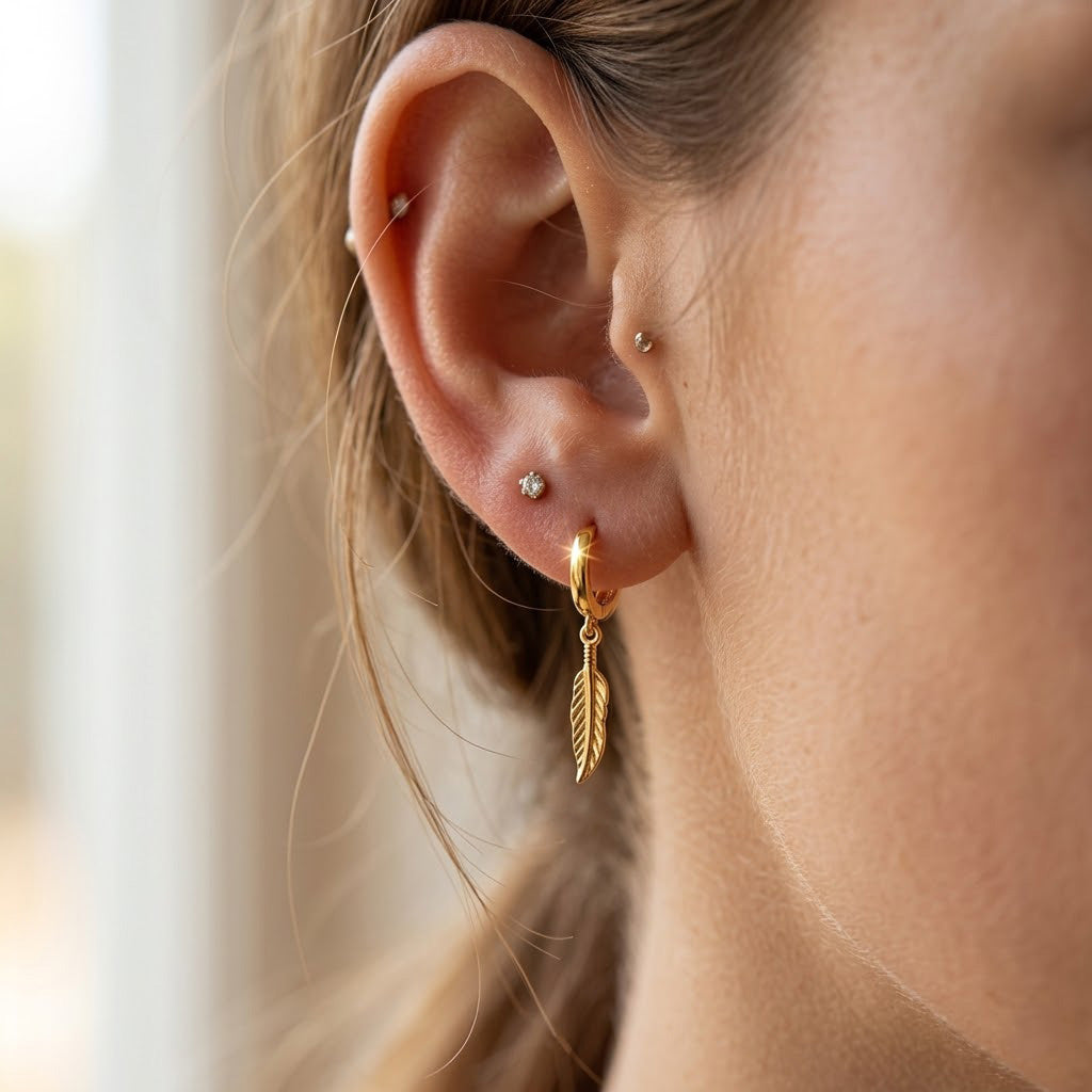 A close-up of a person's ear showcasing a gold huggie hoop earring with a dangling textured feather charm. The person has other small stud earrings on their other ear cartilage and lobe.