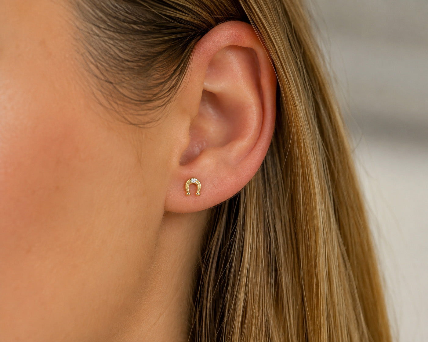A close-up of a woman's ear wearing a small gold horseshoe stud earring.