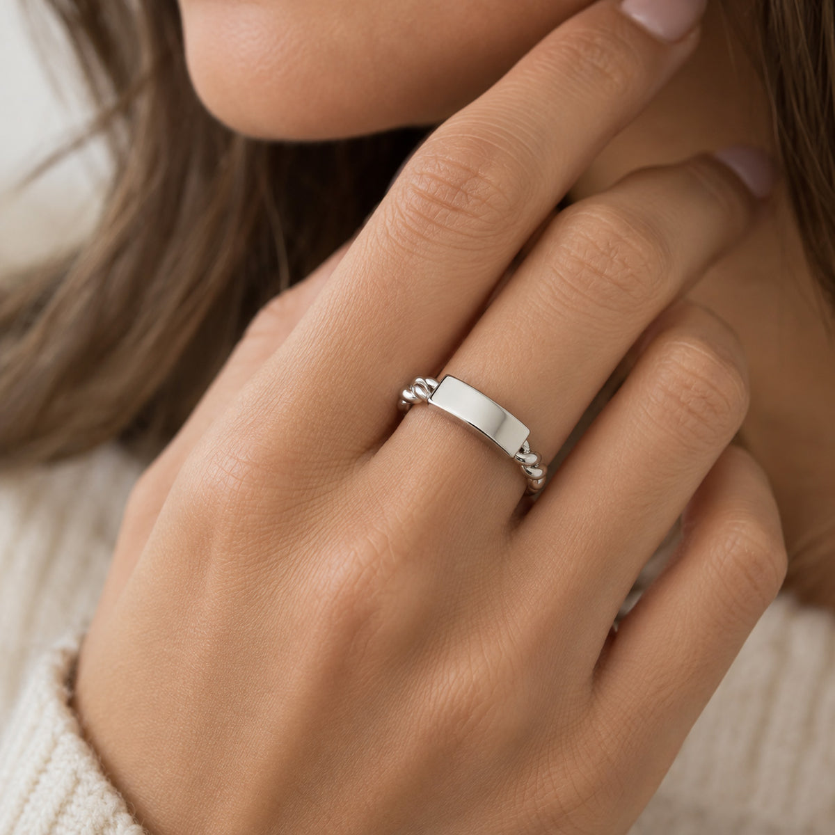 A close-up of a person's hand wearing a polished silver band ring with a small decorative bead detail on one side, set against a soft-focus background of hair and a cream knit sweater.
