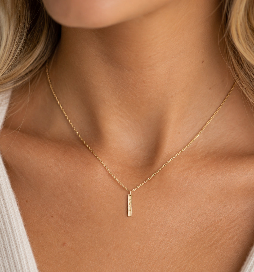 A close-up of a delicate gold chain necklace worn by a model, featuring a small vertical bar pendant with engraved text, set against warm skin tones and a white ribbed top.