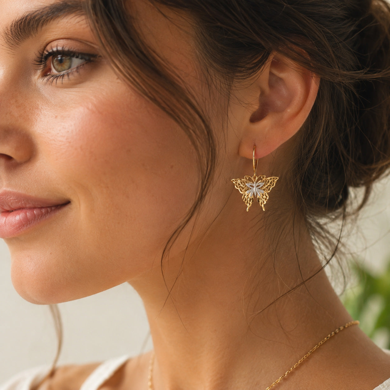 A close-up of a woman wearing a delicate gold butterfly drop earring with a white center, paired with a thin gold chain necklace.