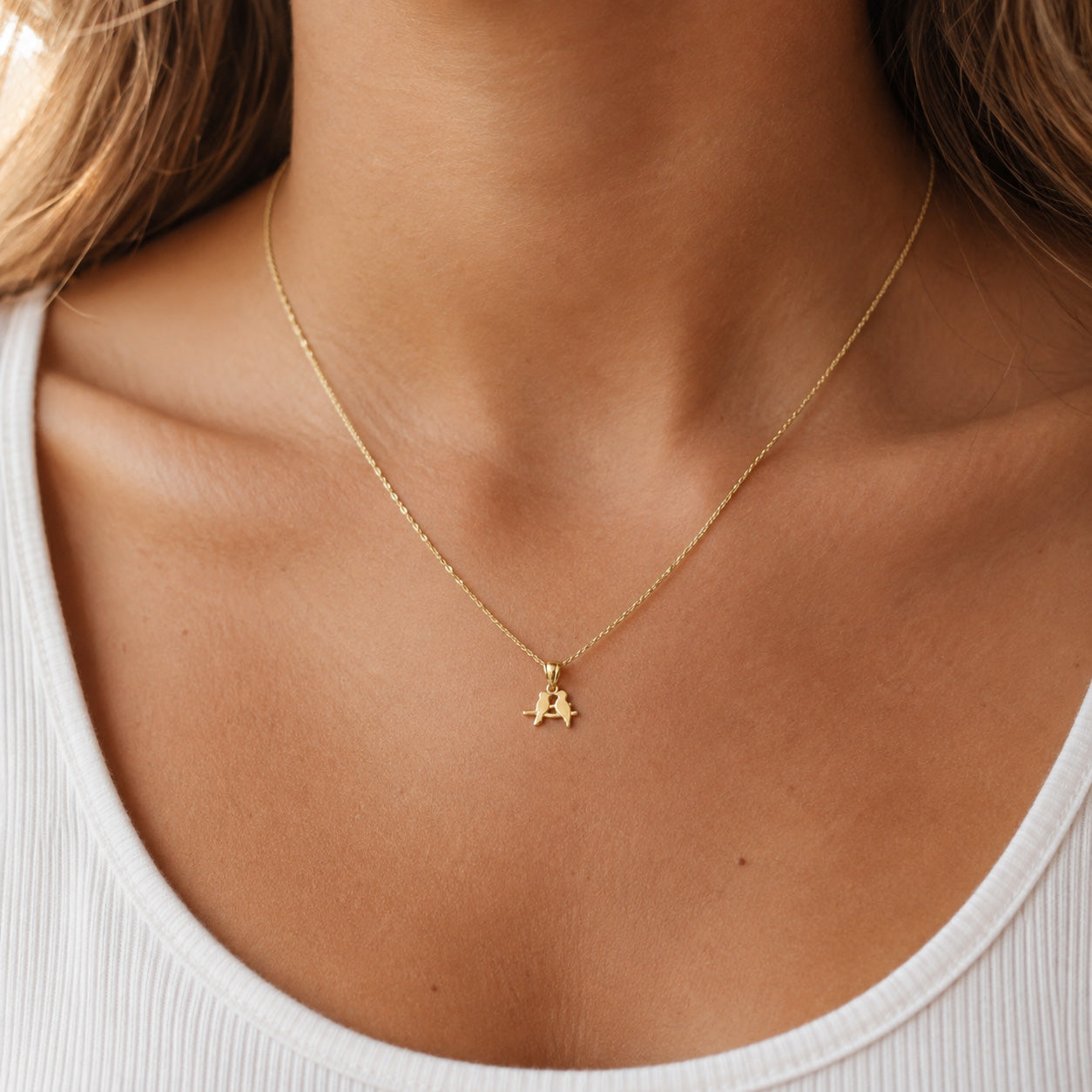 A delicate gold chain necklace with a small letter 'A' pendant, worn on the neck of a model wearing a white ribbed tank top.
