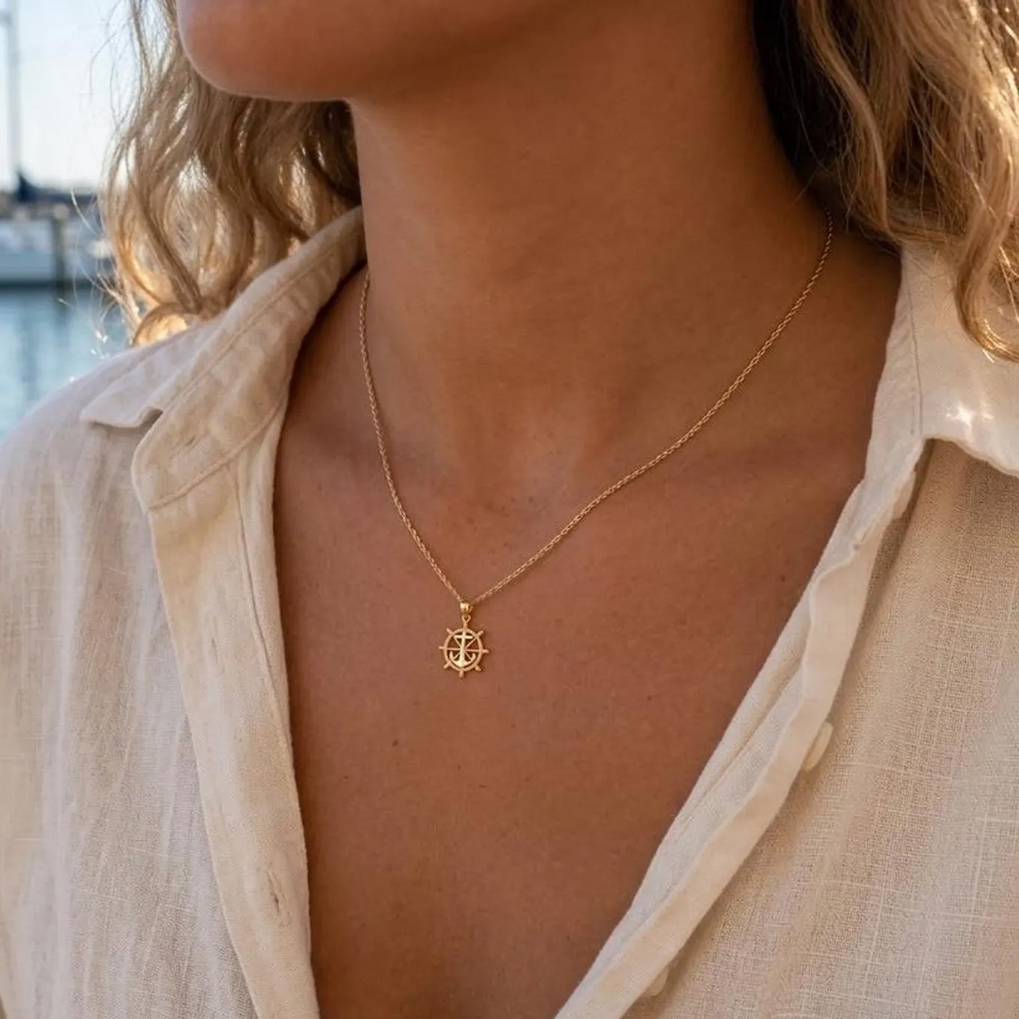Close-up of a person wearing a gold necklace with a pendant, set against a blurred outdoor background.