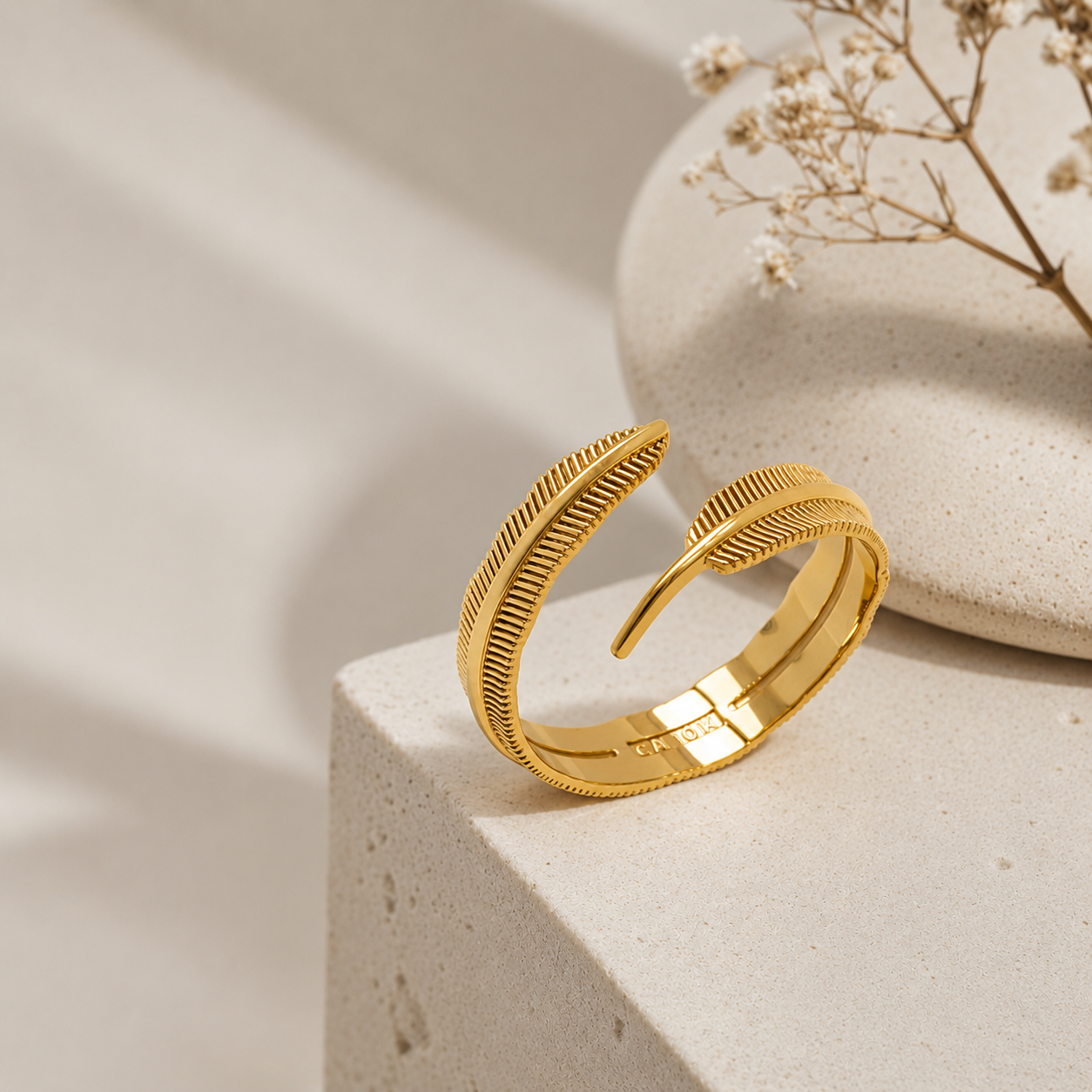 Gold feather-shaped ring on a beige stone surface with dried flowers in the background