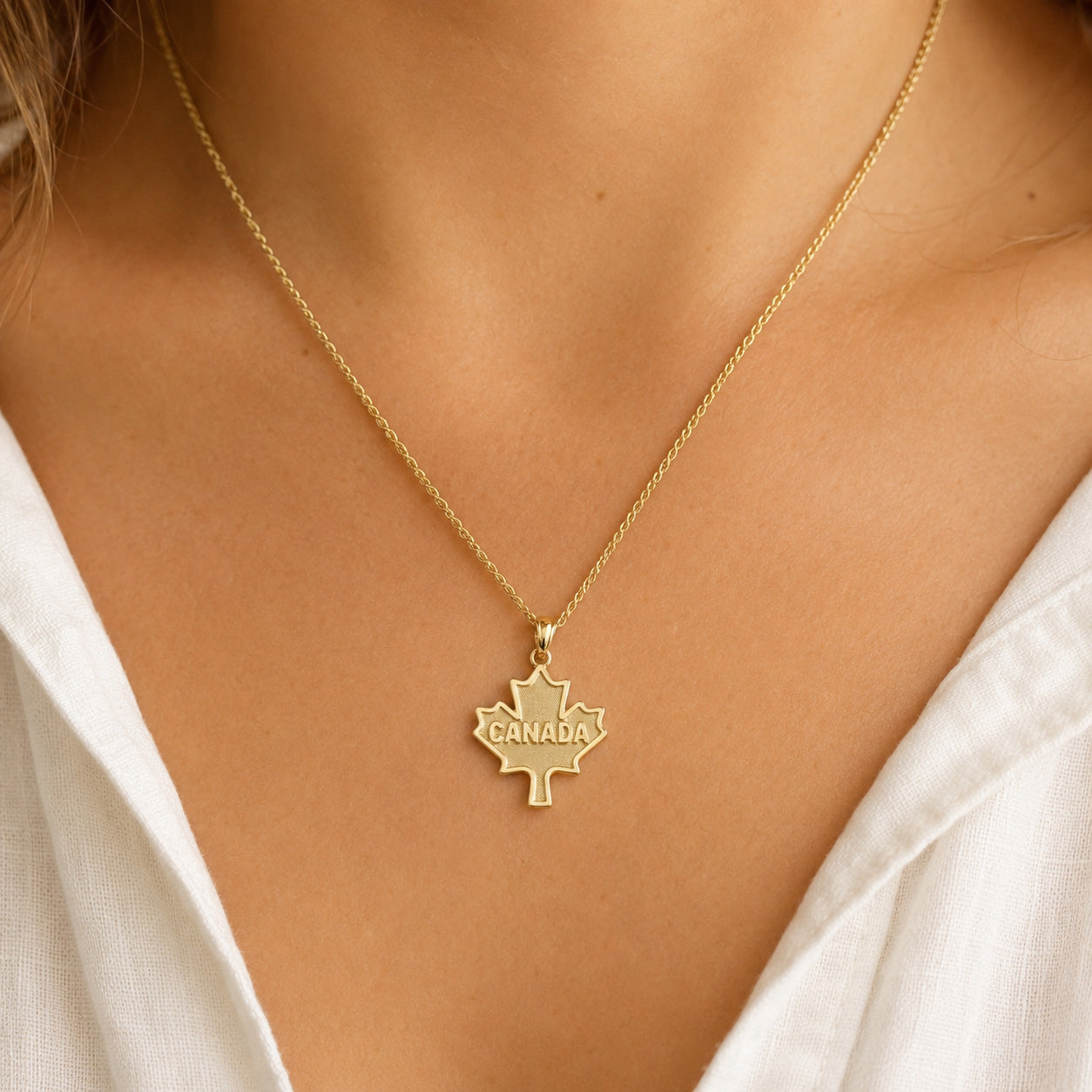 A gold necklace with a maple leaf pendant engraved with the word 'CANADA', worn by a model in a white top.