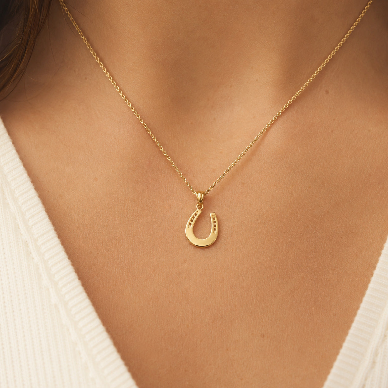 A close-up of a gold horseshoe pendant necklace worn on a model's neck, paired with a white ribbed V-neck top.