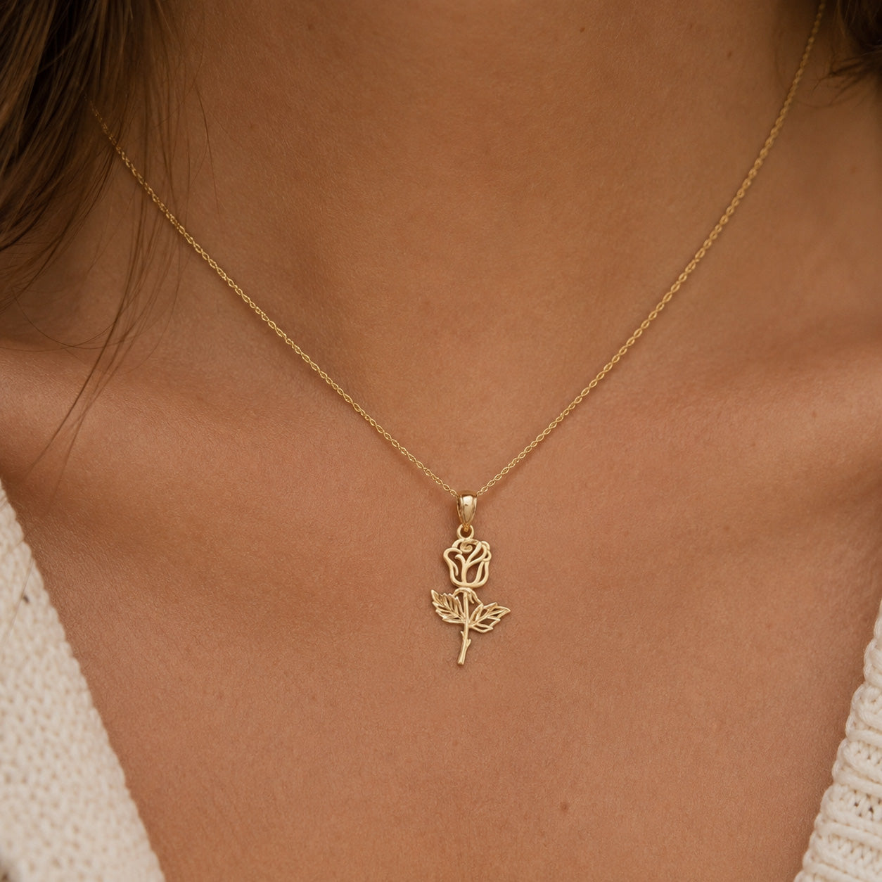 A delicate gold chain necklace with a rose-shaped pendant, worn by a model. The necklace is shown close-up against the model's neck and upper chest, with a cream-colored knit top visible at the edges.