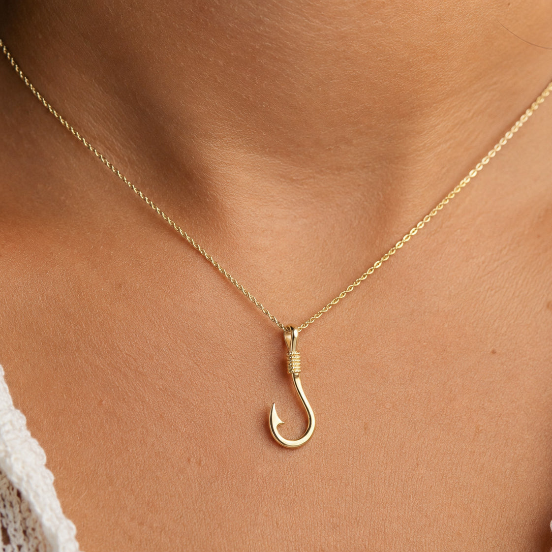 A gold necklace with a hook-shaped pendant worn on a person's neck, shown in a close-up shot with a white lace top visible at the edges.