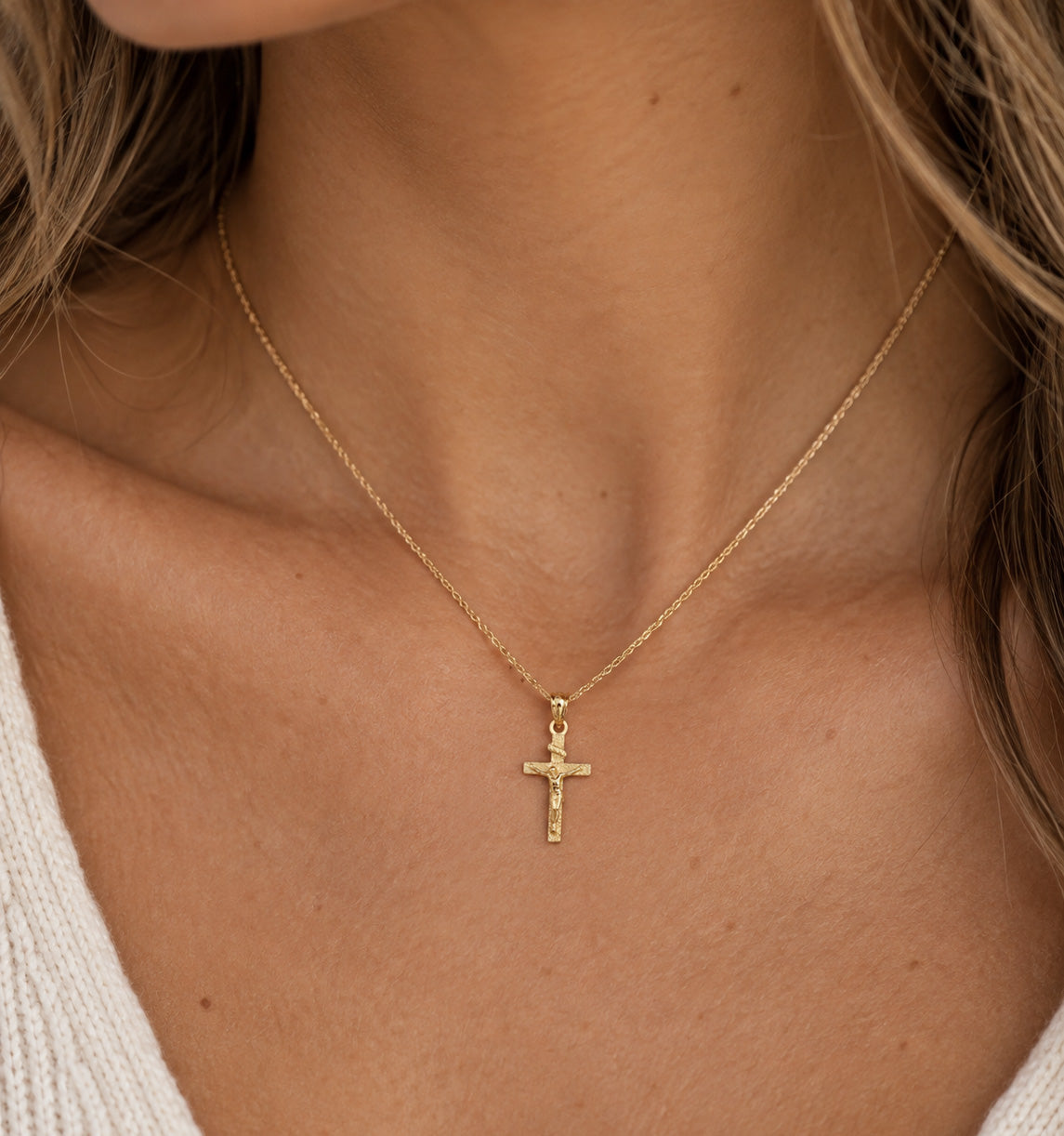 A close-up of a model's neck and chest wearing a delicate gold chain necklace with a small cross pendant.