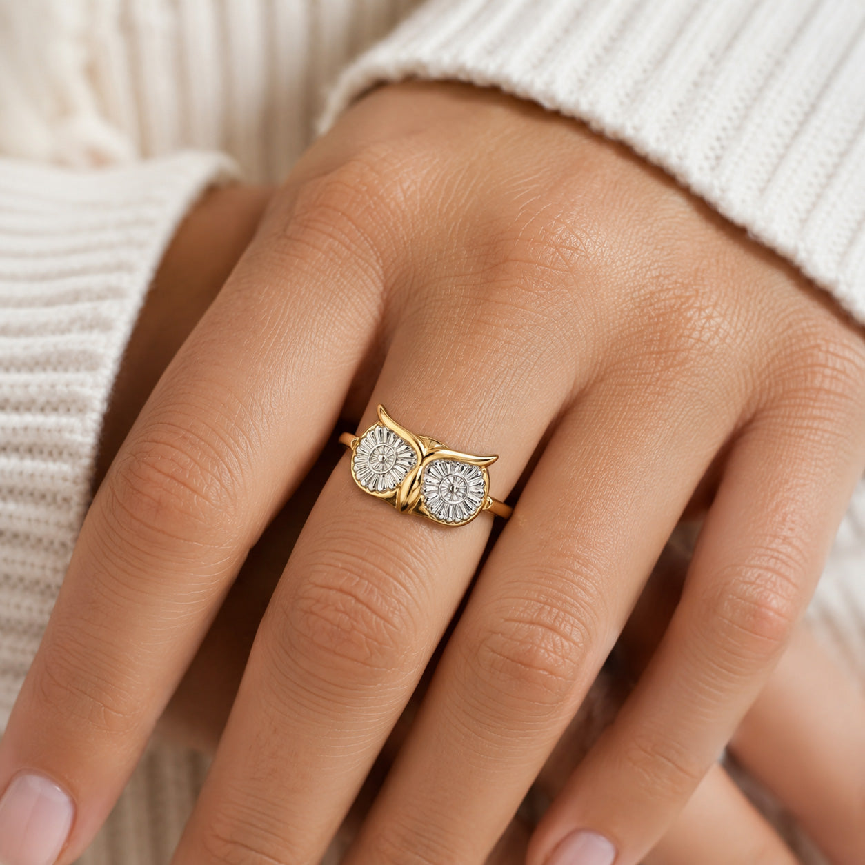A gold owl-shaped ring with two round clear gemstones on a person's ring finger, shown close-up against a white ribbed sweater.