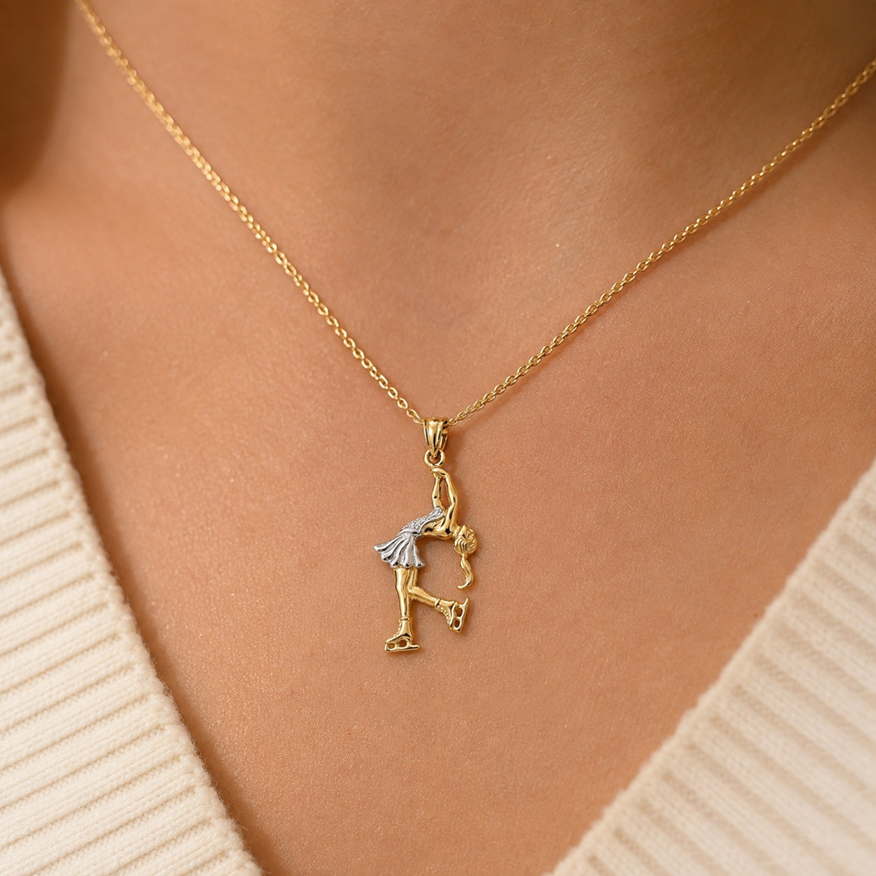 A gold chain necklace with a dangling figure pendant worn on a person's neck, shown against a cream ribbed sweater.