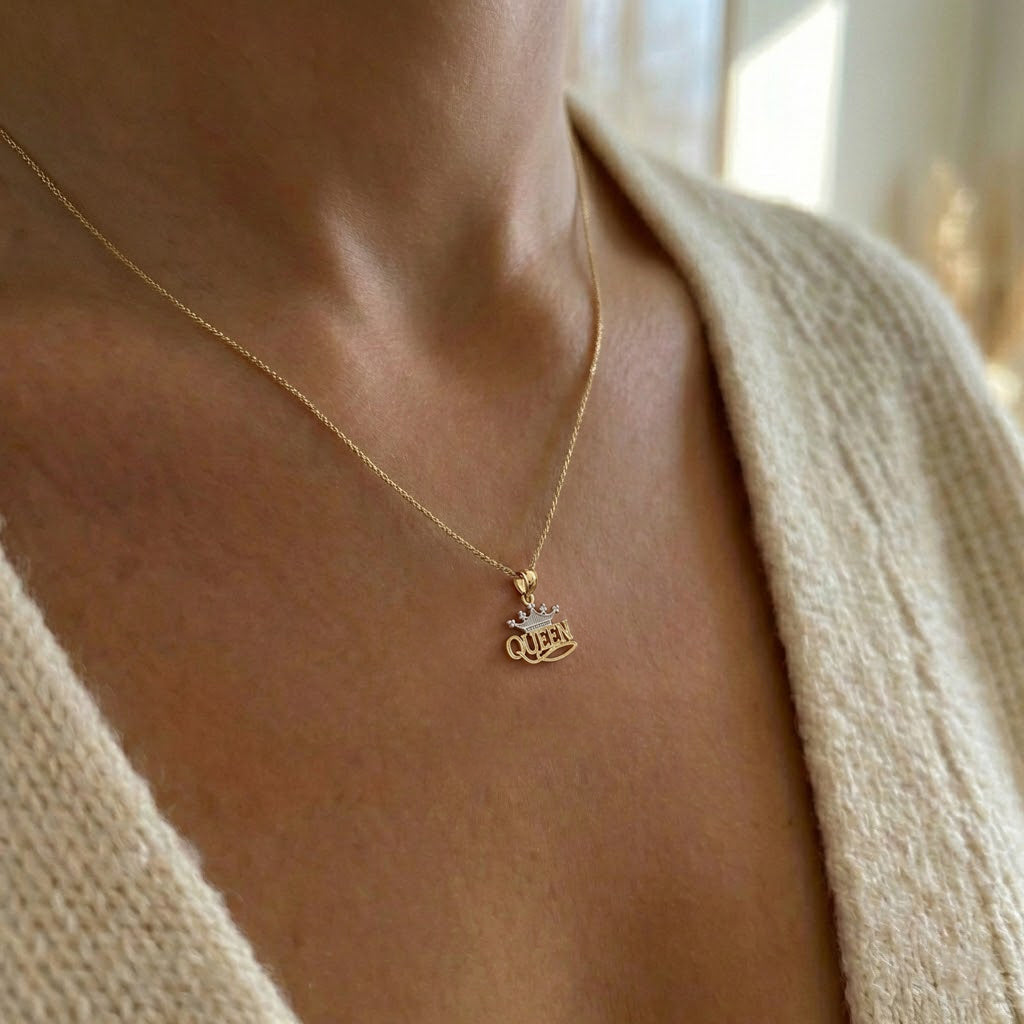 A close-up of a person wearing a delicate gold chain necklace with a small script pendant spelling 'Queen', styled with a cream knit cardigan.