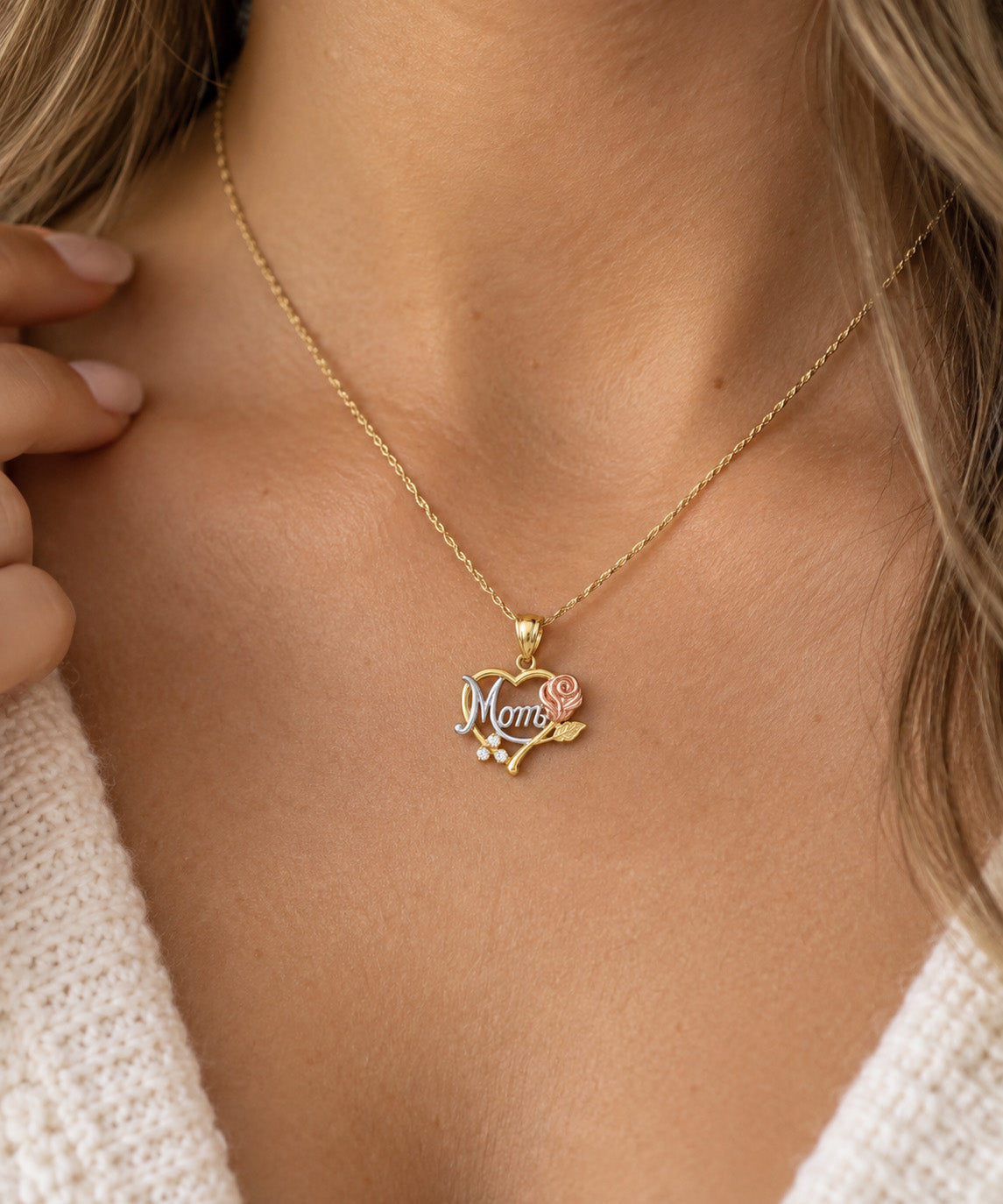 A close-up of a woman's neck and chest wearing a delicate gold chain necklace with a heart-shaped pendant. The pendant features the word 'Mom' in silver script, a small pink rose, and tiny white pearl-like accents.