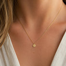 A delicate gold chain necklace with a small, round, textured gold pendant worn by a model. The necklace is shown close-up against the model's neck and collarbone, wearing a white top.