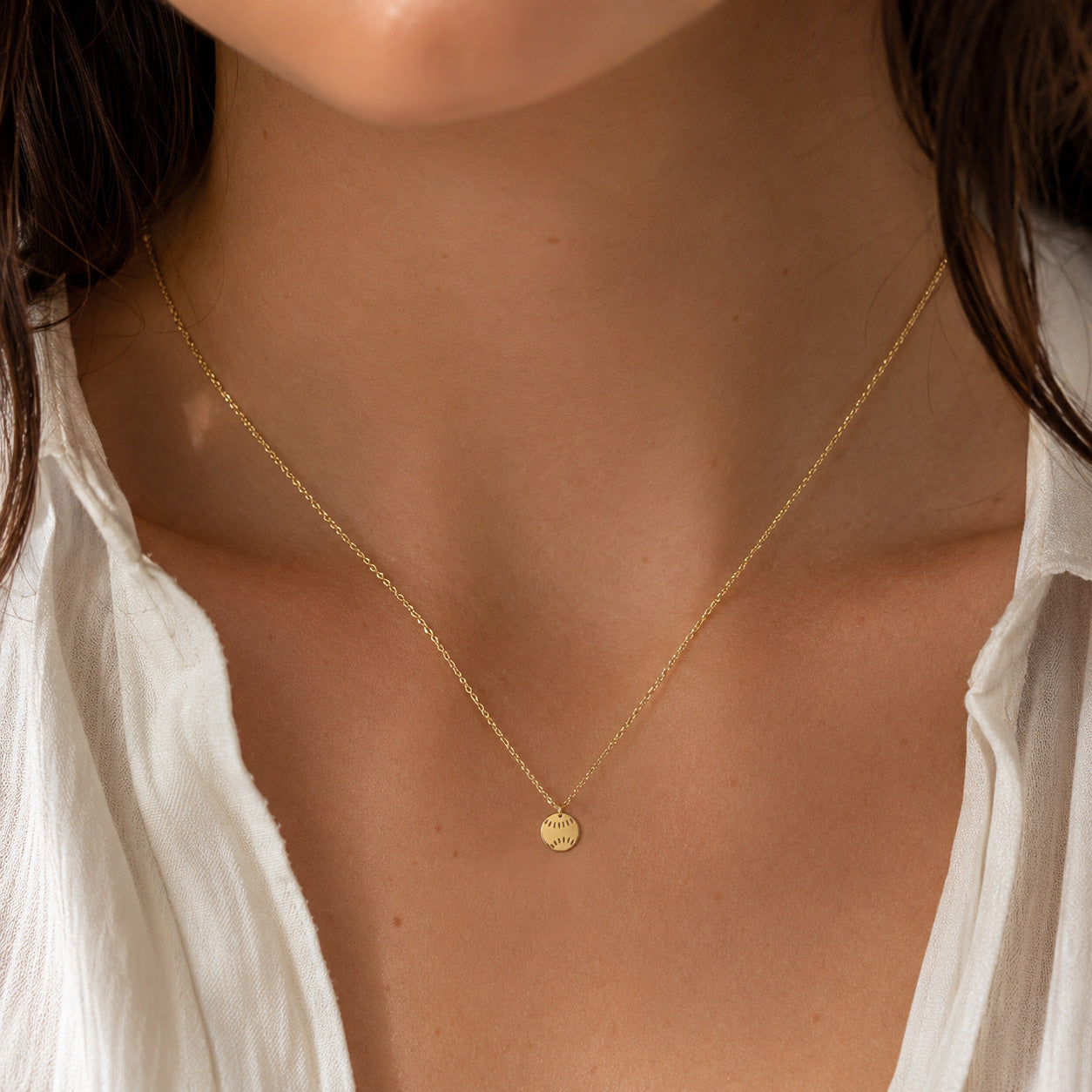 A delicate gold chain necklace with a small round gold disc pendant worn by a model. The model is wearing a white top, and the background is softly blurred.