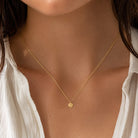 A delicate gold chain necklace with a small round gold disc pendant worn by a model. The model is wearing a white top, and the background is softly blurred.