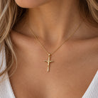 A close-up of a woman wearing a delicate gold chain necklace with a small gold crucifix pendant, styled with a white ribbed top.