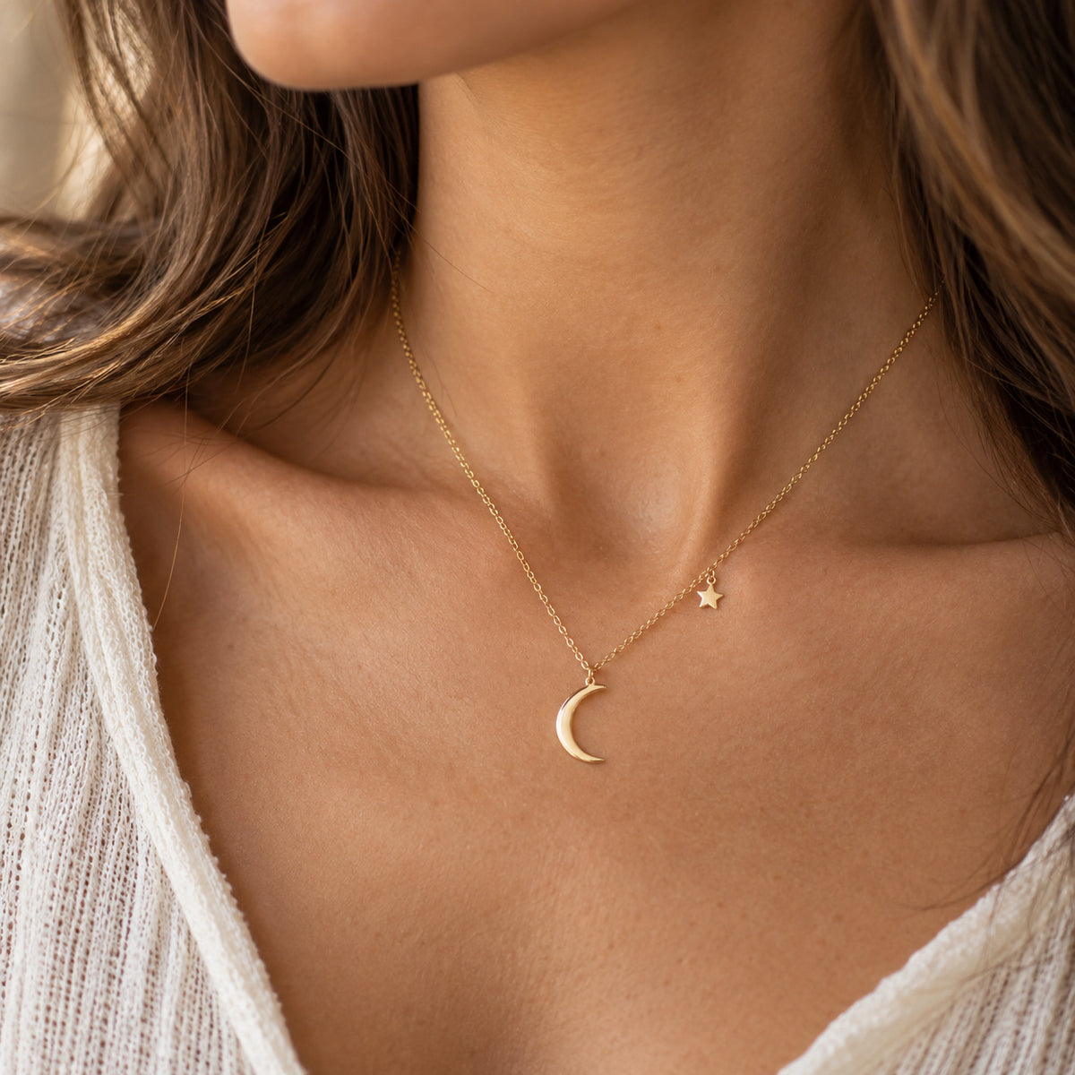 A close-up of a woman's neck and chest wearing a delicate gold necklace with a crescent moon and a small star pendant.