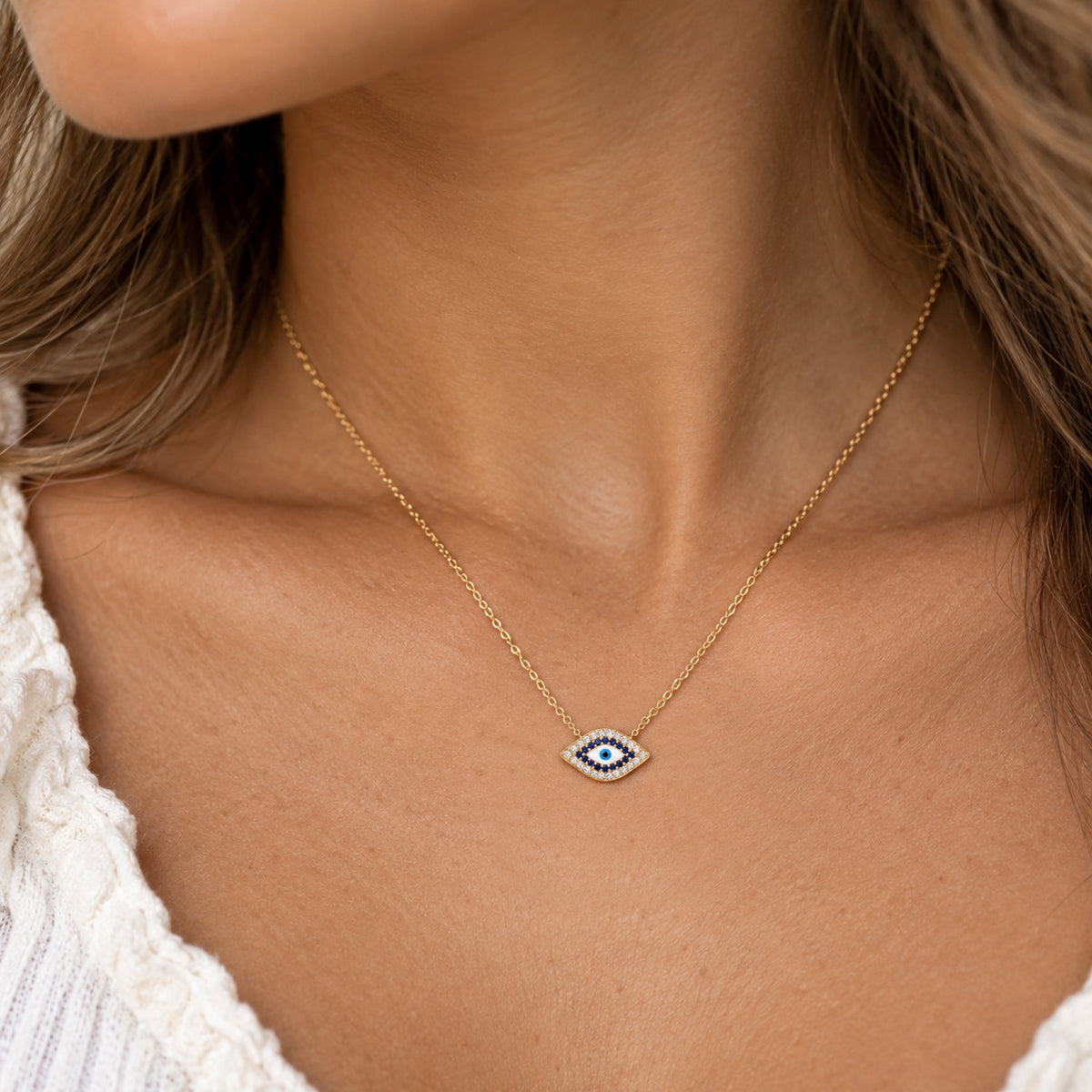 A close-up of a woman's neck and chest wearing a delicate gold chain necklace with a small evil eye pendant. The pendant features a white background with a blue center and small black details, surrounded by a silver border.