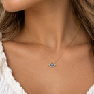 A close-up of a woman's neck and chest wearing a delicate gold chain necklace with a small evil eye pendant. The pendant features a white background with a blue center and small black details, surrounded by a silver border.