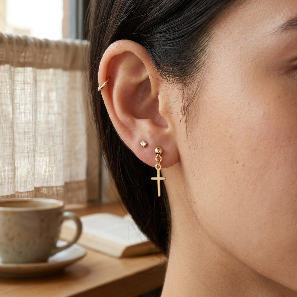 Close-up of a person wearing gold cross earrings with a blurred background of a cup and book on a table.