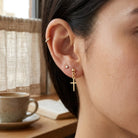 Close-up of a person wearing gold cross earrings with a blurred background of a cup and book on a table.