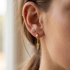 A close-up of a person's ear showcasing a gold huggie hoop earring with a dangling textured feather charm. The person has other small stud earrings on their other ear cartilage and lobe.