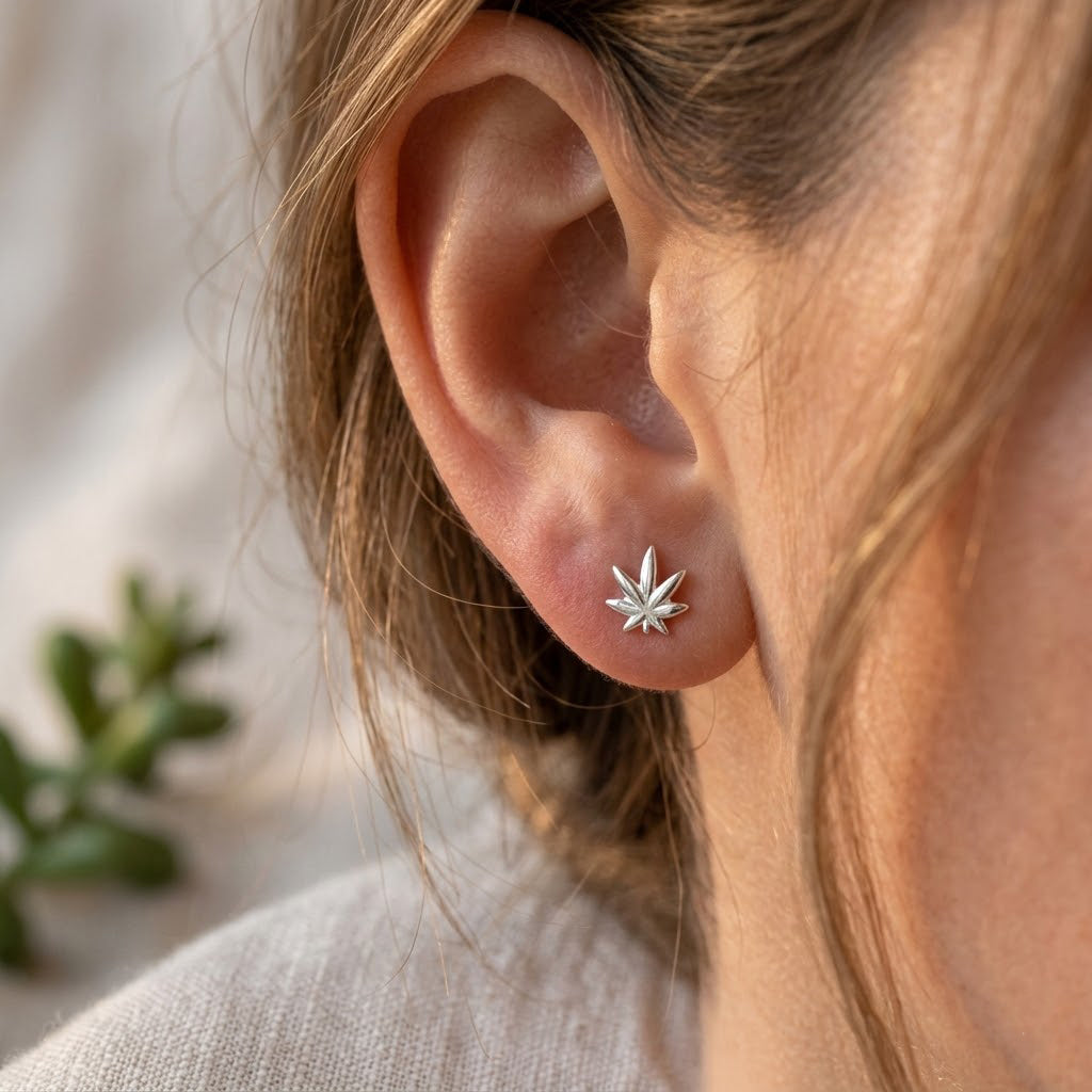 A close-up of a person's ear wearing a small silver stud earring shaped like a six-pointed star or leaf, with soft-focus hair and a neutral background.