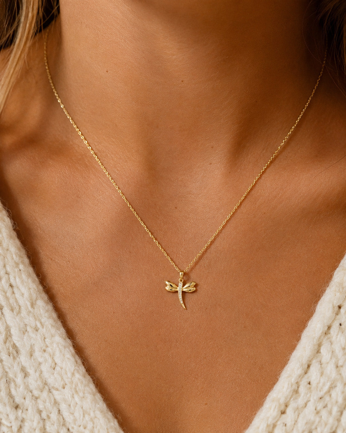 A delicate gold chain necklace with a small dragonfly pendant, worn by a model in a close-up shot. The model is wearing a cream-colored knit sweater.