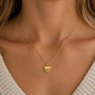 [group: large] A close-up of a model wearing a delicate gold chain necklace with a textured, ridged heart-shaped pendant.