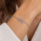 [group:sco]   A silver chain bracelet featuring a dangling zodiac sign 'Scorpio' charm paved with small clear stones, shown on a plain white background.A silver-toned Pisces zodiac sign bracelet with small clear stones, shown worn on a person's wrist.