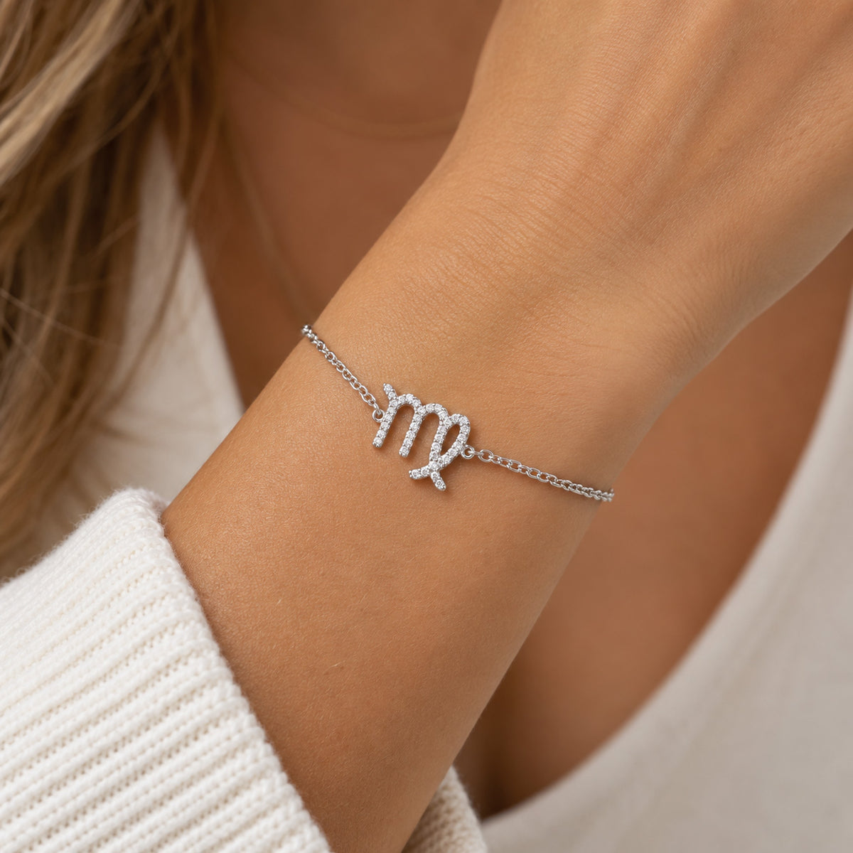 [group: vir] silver virgo braceletA close-up of a woman's wrist wearing a delicate silver chain bracelet with a zodiac sign charm, likely representing Libra, set with small clear stones. She is wearing a white knit top.