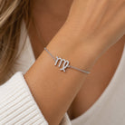 [group: vir] silver virgo braceletA close-up of a woman's wrist wearing a delicate silver chain bracelet with a zodiac sign charm, likely representing Libra, set with small clear stones. She is wearing a white knit top.