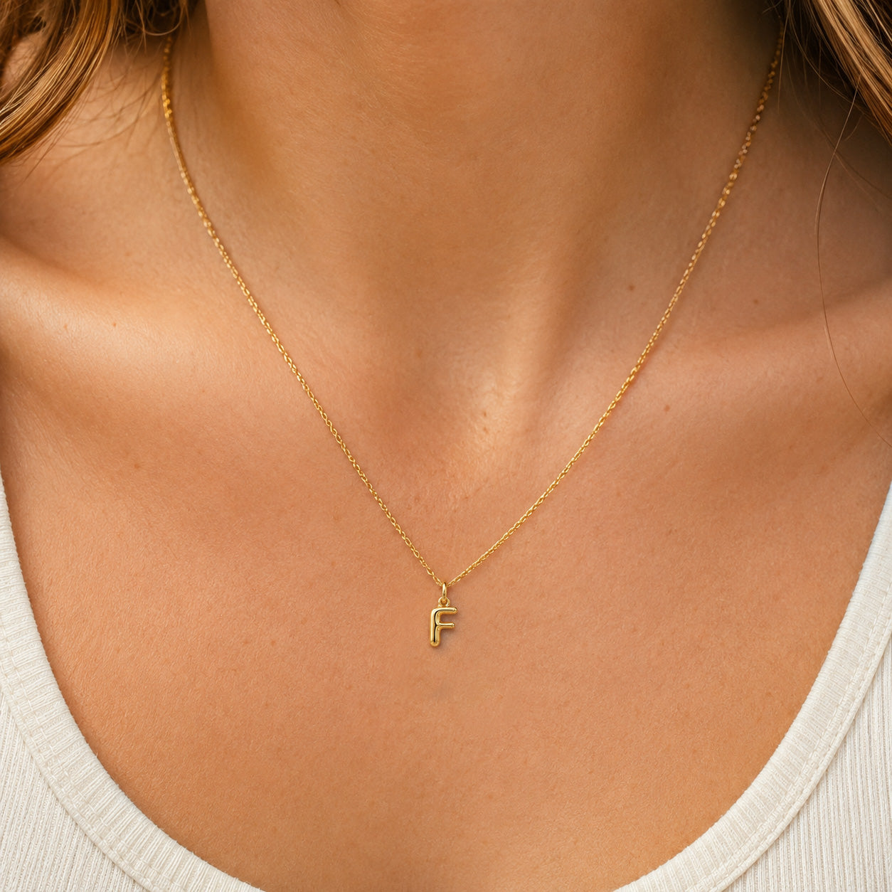 [group: f-10k-gold]A delicate gold chain necklace with a small letter 'F' initial pendant, worn on the neck of a model wearing a white ribbed top.