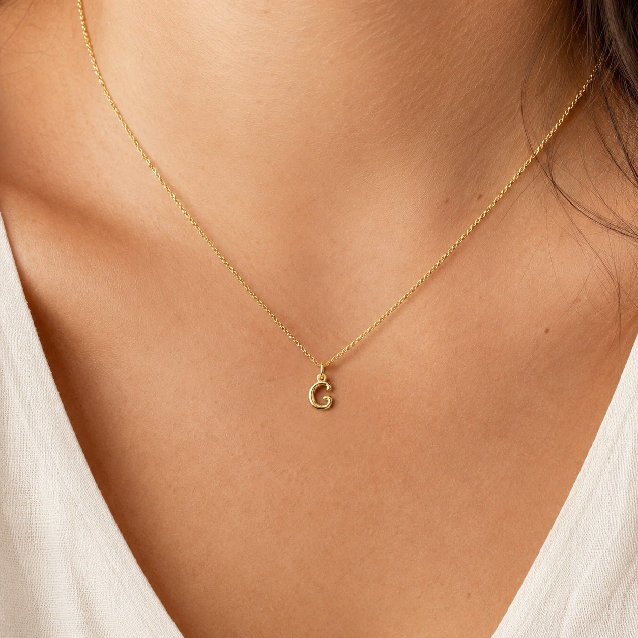 [group: g-10k-gold]A delicate gold chain necklace with a small lowercase letter 'c' initial pendant, shown worn on a model's neck over a white V-neck top.
