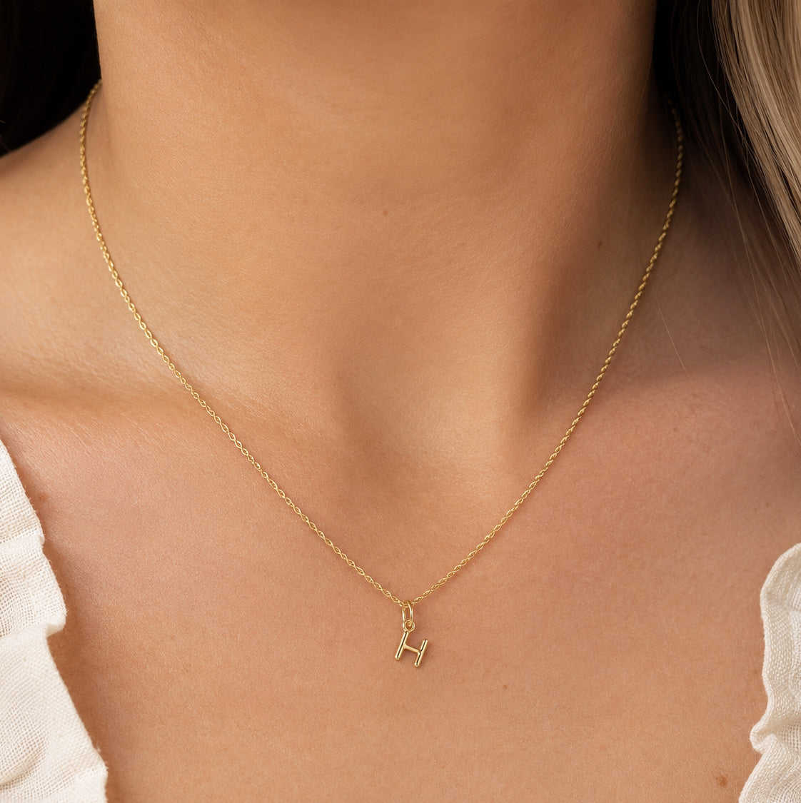 [group: h-10k-gold]A delicate gold chain necklace with a small initial 'H' pendant, worn on the neck of a model.