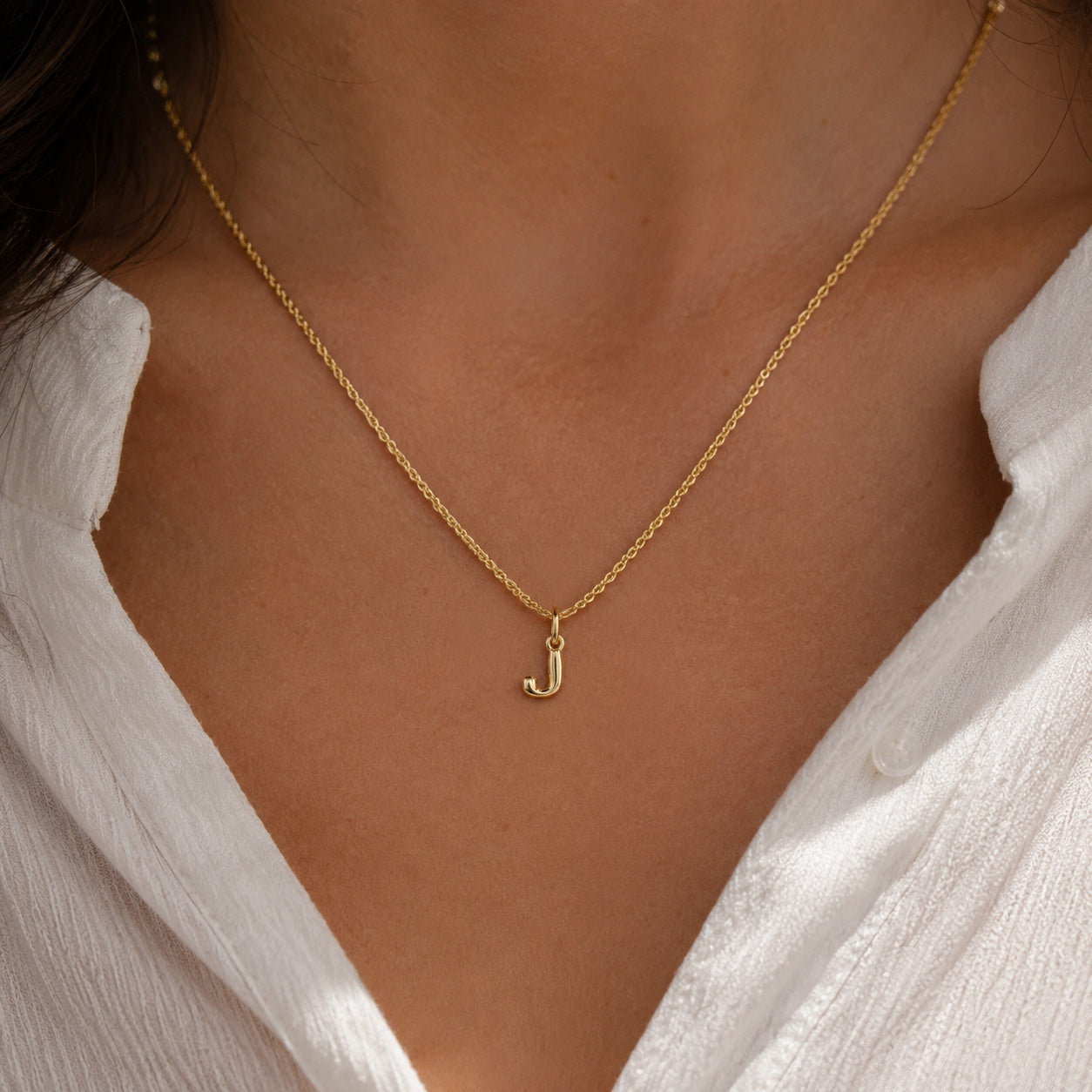 [group: j-10k-gold]A delicate gold chain necklace with a small lowercase letter 'j' pendant, worn on a model's neck over a white collared shirt.