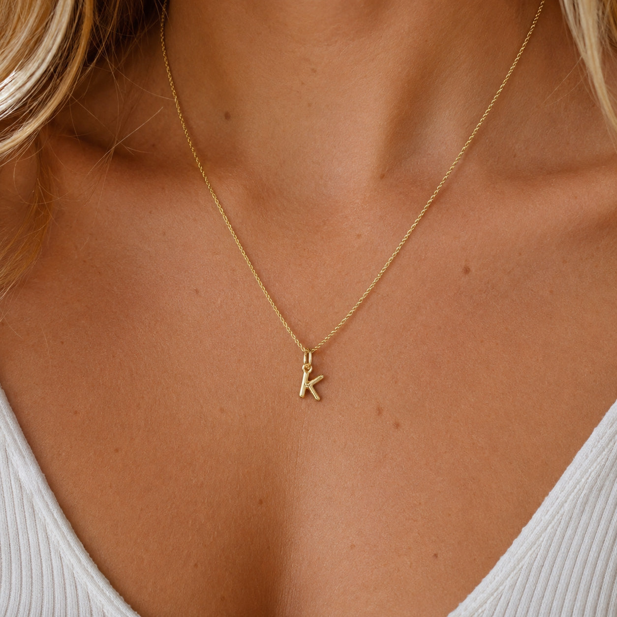[group: k-10k-gold]A close-up of a model's neck and chest wearing a delicate gold chain necklace with a small initial pendant spelling 'K'.