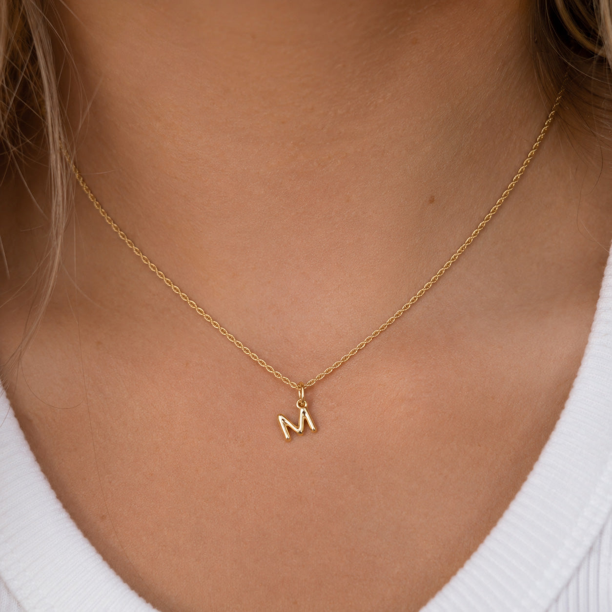 [group: m-10k-gold]A delicate gold chain necklace with a small lowercase letter 'm' pendant, worn on a model's neck over a white ribbed top.