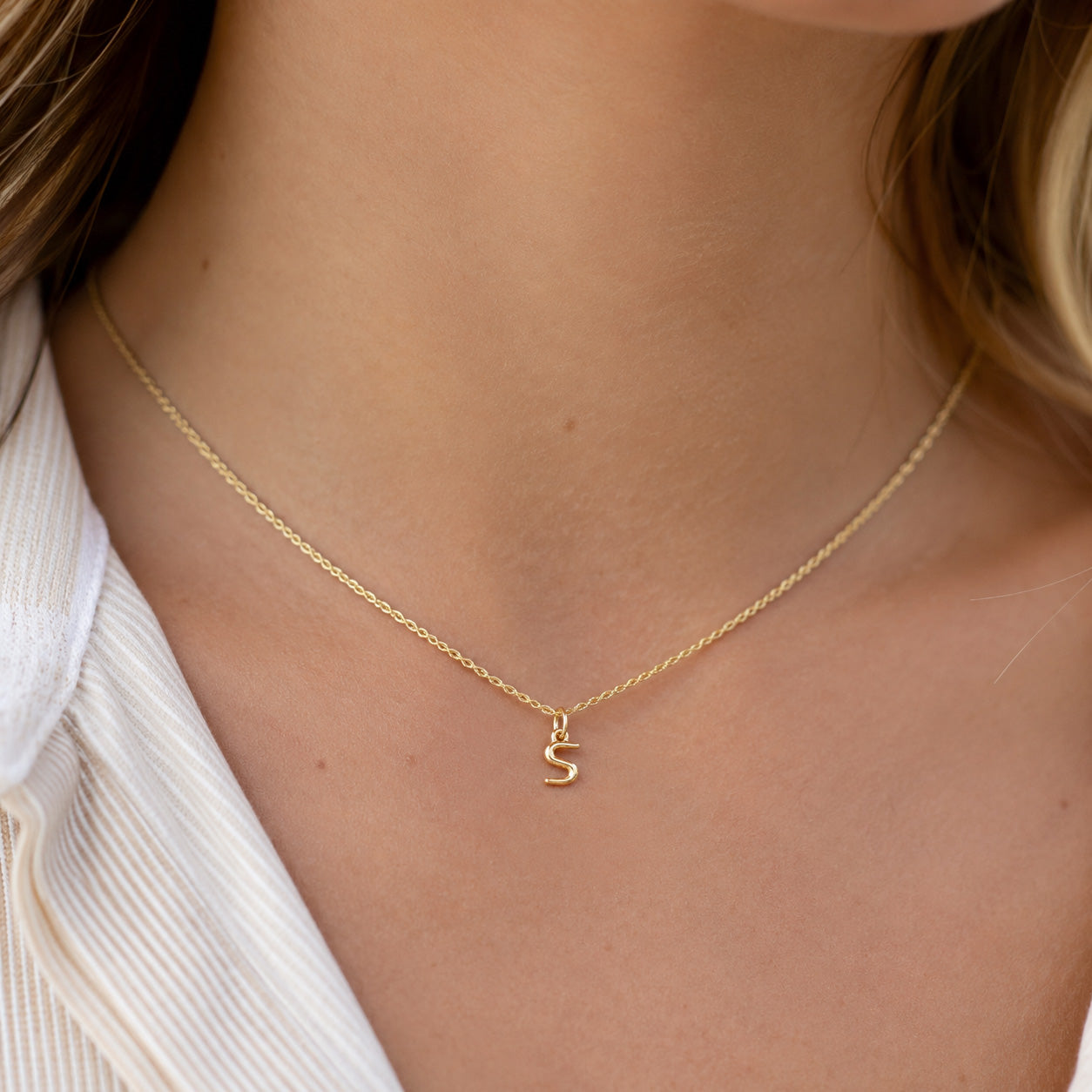 [group: s-10k-gold]A delicate gold chain necklace with a small initial 'S' pendant, worn on a model's neck over a white ribbed top.