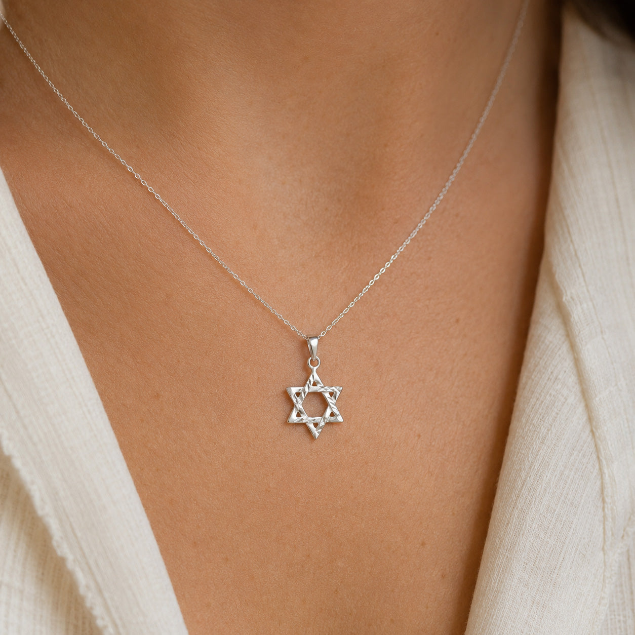 A close-up of a delicate silver chain necklace with a Star of David pendant, worn by a person in a cream-colored top.