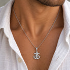 A silver anchor pendant necklace worn by a man, shown close-up against his neck and white shirt.