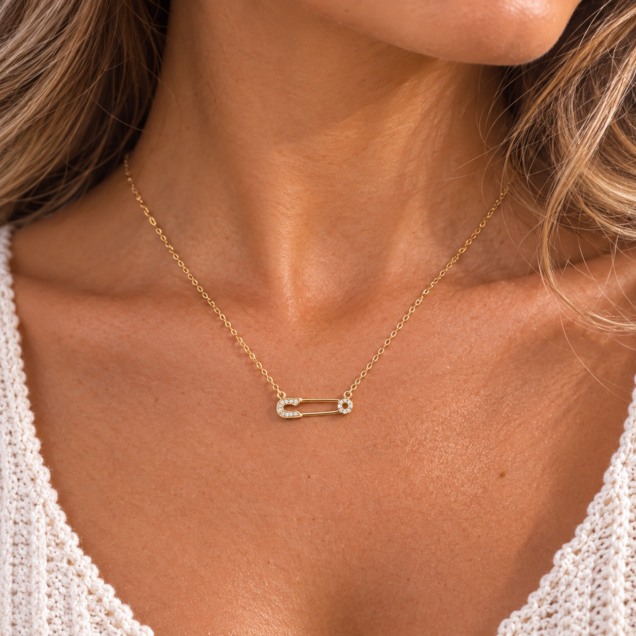 [group: gold-plated]A close-up of a woman's neck and chest wearing a delicate gold safety-pin necklace with small clear stones on the front, paired with a white knit top.