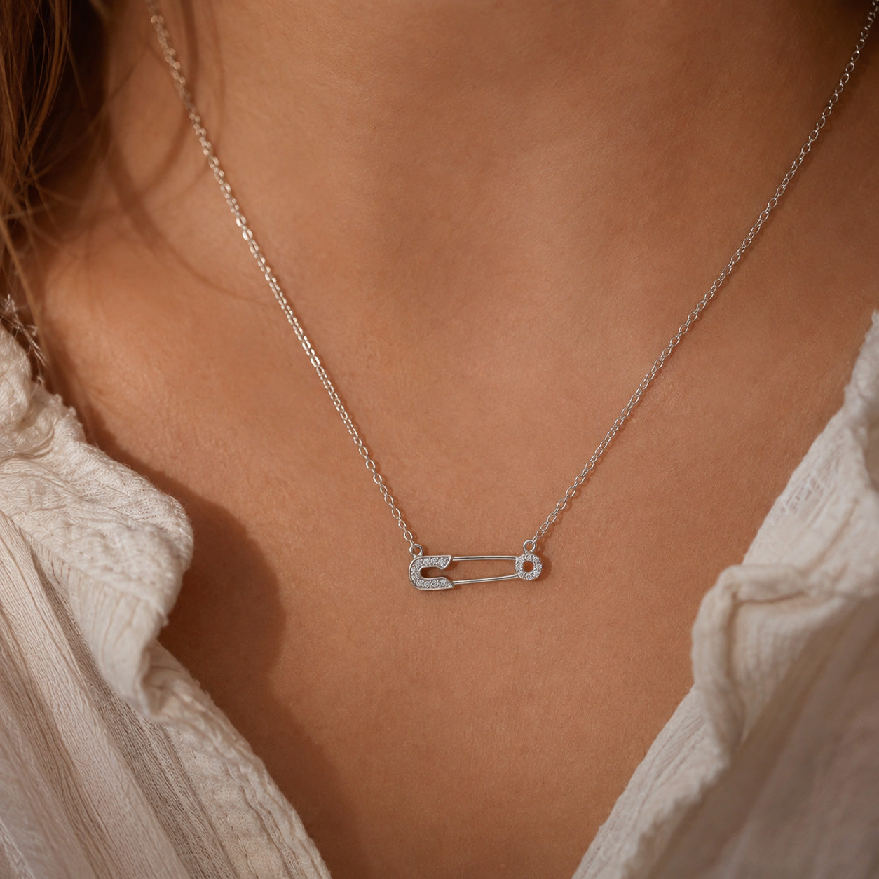 [group: silver]A silver safety pin necklace with small clear stones, worn on a model's neck over a white textured blouse.