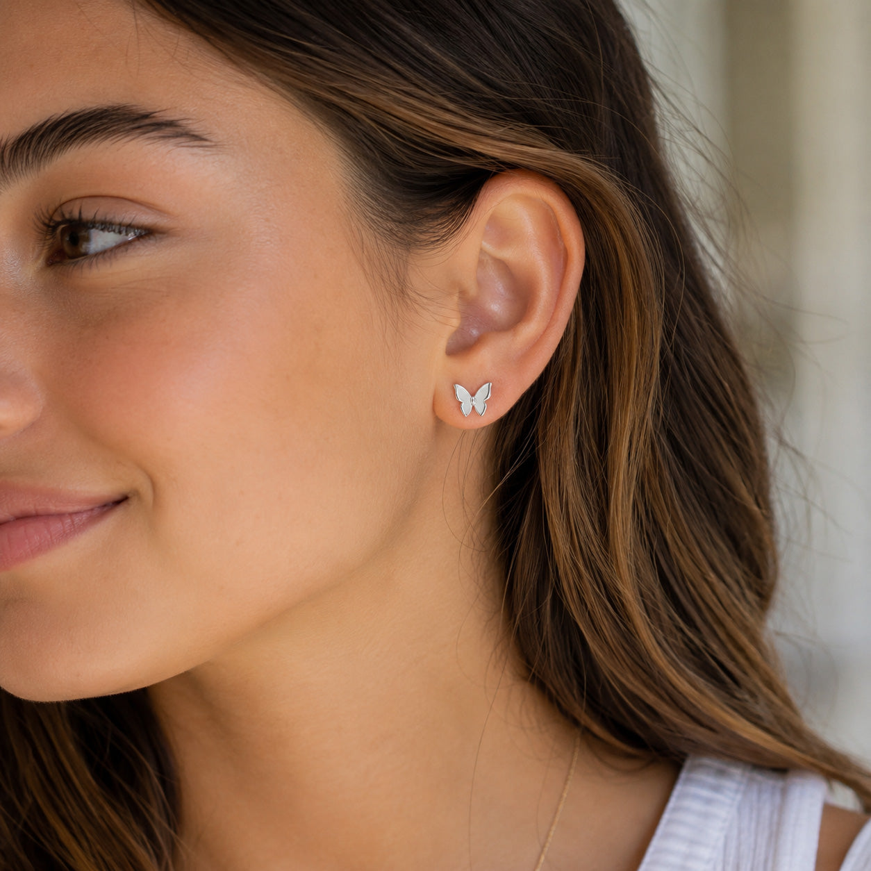 A close-up of a woman's ear wearing a small silver butterfly stud earring, with her brown hair and skin visible against a softly blurred light background.