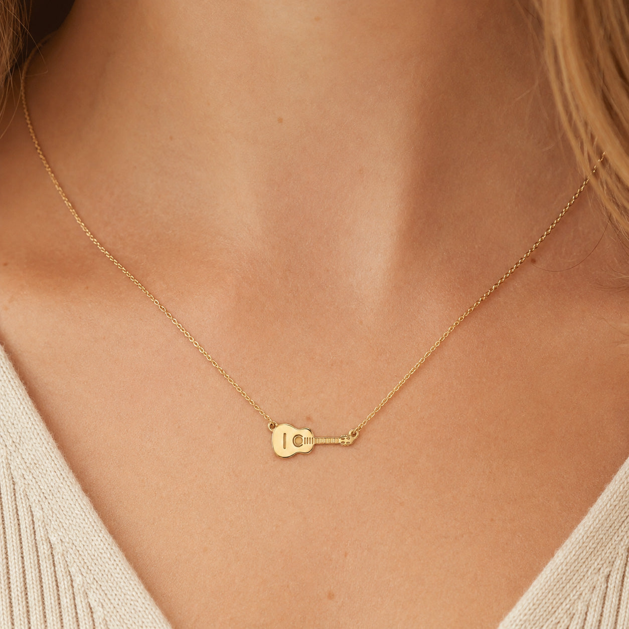 A delicate gold necklace with a small guitar-shaped pendant, worn on a model's neck over a cream-colored knit top.