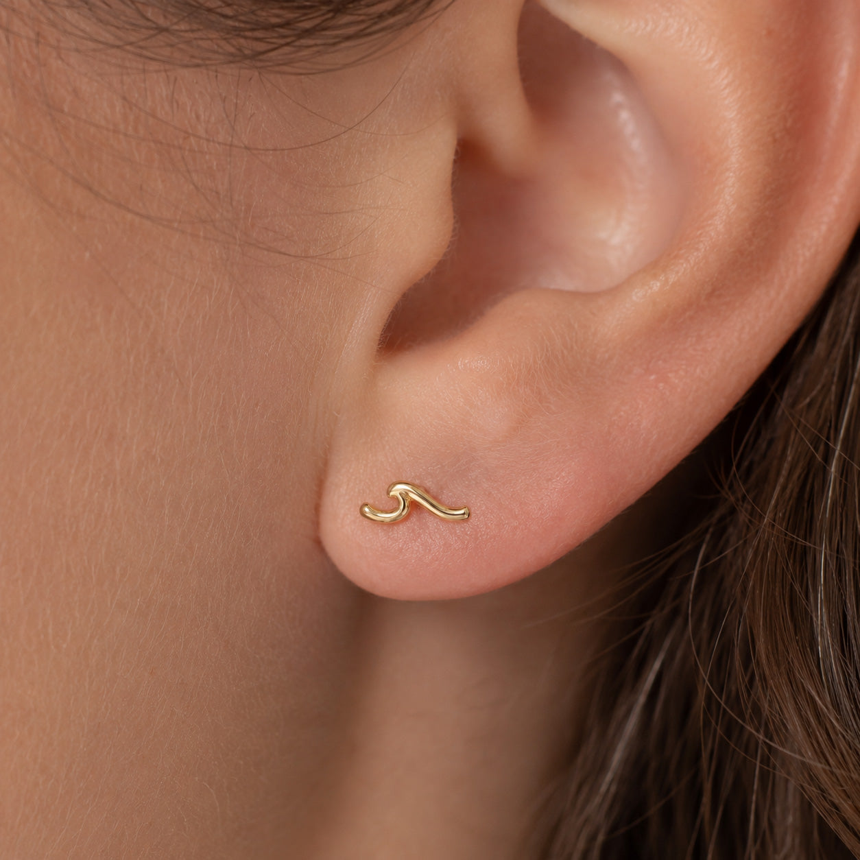 A close-up of a person's ear wearing a small, delicate gold wavy stud earring.